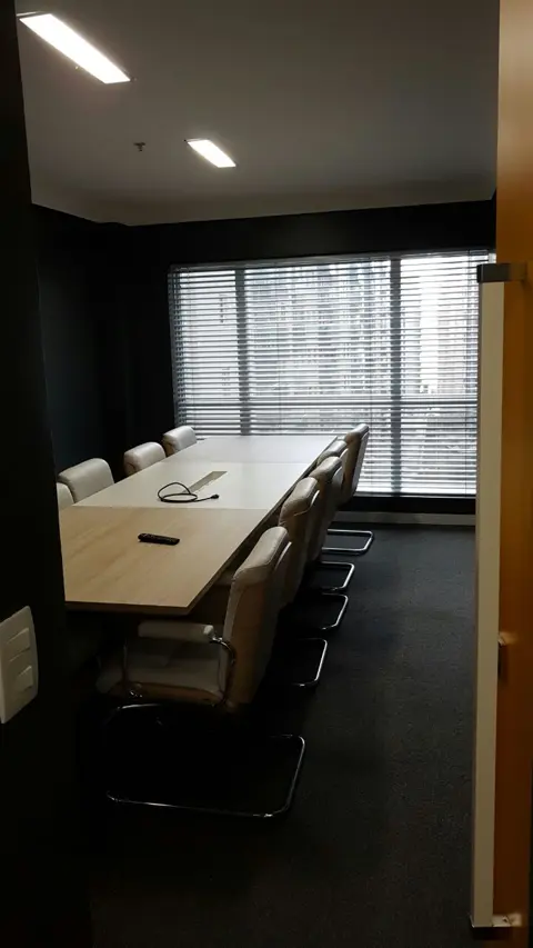 Mesa de trabalho compartilhada no coworking, com cadeiras e janela com persianas